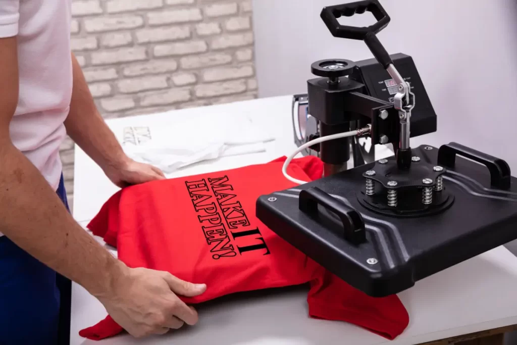 Uma pessoa posiciona uma camiseta vermelha com a frase preta "MAKE IT HAPPEN!" em uma prensa térmica preta ao lado.