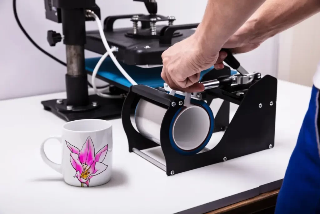 A mão de um homem fecha a alavanca de uma prensa térmica cilíndrica preta, pressionando uma caneca branca com uma estampa de tulipa rosa e amarela contra o mandril aquecido. Uma segunda caneca estampada idêntica está sobre a mesa branca à esquerda.