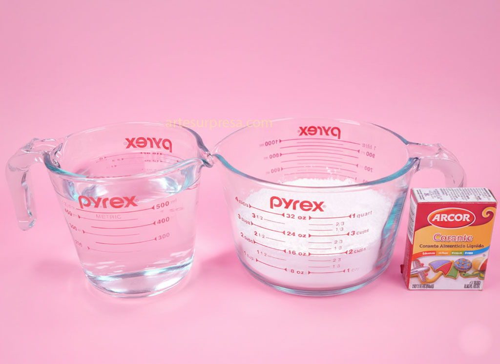 Dois copos medidores de vidro Pyrex sobre um fundo rosa. O copo à esquerda contém água, e o copo à direita contém um pó branco (provavelmente farinha). Ao lado direito do copo maior, há uma caixa de corante alimentício líquido da marca Arcor.
